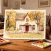 May include: Watercolour painting of a white house with red trim, a red door, and a white picket fence. The house is surrounded by trees with yellow and orange leaves, suggesting an autumn scene. The painting is on a wooden table with paint palettes and brushes.