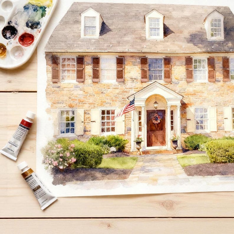May include: Watercolour painting of a two-storey house with a stone facade, brown shutters, and a front door with a wreath. An American flag hangs near the entrance. Paint tubes and a palette with watercolours are in the foreground.