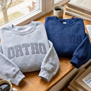 Könnte beinhalten: Zwei Rundhals-Sweatshirts, eines in Hellgrau und eines in Marineblau, jeweils mit dem Wort ORTHO in einer College-Schriftart auf der Brust bestickt. Auf dem linken Ärmel jedes Sweatshirts ist der Name Emma in Schreibschrift eingestickt.