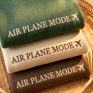 Puede incluir: Tres mantas plegadas, apiladas, en tonos verde, beige y marr&oacute;n. Cada manta tiene bordada la frase "AIR PLANE MODE" en blanco, junto con un peque&ntilde;o gr&aacute;fico de avi&oacute;n. Disposici&oacute;n ordenada.