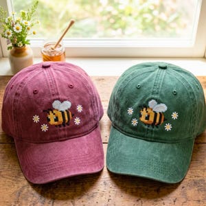 May include: Two baseball caps, one in maroon and one in forest green, each featuring an embroidered pixel-art style bee surrounded by small white daisy flowers. Both caps have a structured crown, a curved brim, and a button on top.