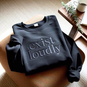 May include: A black sweatshirt with the embroidered phrase "exist loudly" in a lighter gray color. The sweatshirt is folded on a patterned chair, with a wooden side table and a mug visible in the background.