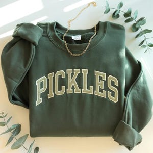 以下が含まれることがあります： 「PICKLES」の文字がライトグリーンで刺繍された、カレッジスタイルのダークグリーンのスウェットシャツ。ゴールドのチェーンネックレスがスウェットシャツの上に置かれています。袖は折り返され、背景には緑の小枝があります。