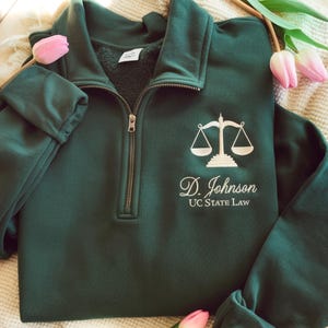May include: A dark green quarter-zip sweatshirt with a white embroidered scales of justice logo and the name "D. Johnson" and "UC STATE LAW" below. The sweatshirt has a fleece lining and a metal zipper.