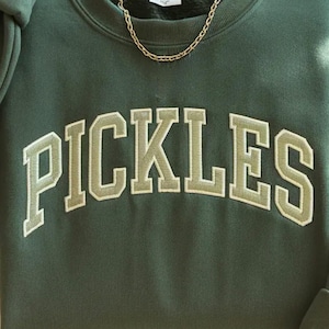 以下が含まれることがあります： 「PICKLES」の文字が大きくアーチ状に配置された、白の縁取りのある明るい緑色の文字が特徴の濃い緑色のスウェットシャツ。上部にはゴールドのチェーンネックレスが見えます。スウェットシャツは柔らかい素材で作られています。