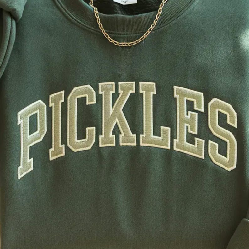 以下が含まれることがあります： 「PICKLES」の文字が大きくアーチ状に配置された、白の縁取りのある明るい緑色の文字が特徴の濃い緑色のスウェットシャツ。上部にはゴールドのチェーンネックレスが見えます。スウェットシャツは柔らかい素材で作られています。