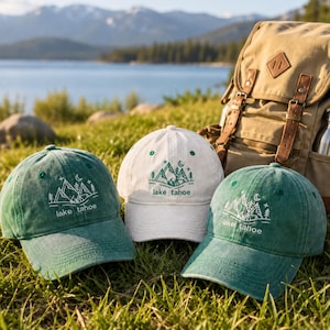 May include: Two green baseball caps and one white baseball cap, each featuring an embroidered mountain landscape design with pine trees, a crescent moon, and the text lake tahoe in a simple line art style, positioned on a grassy surface.