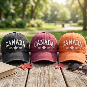 May include: Three baseball caps in black, maroon, and orange, each featuring white embroidered text that reads CANADA EST. 1867 with a maple leaf icon. The caps have a structured crown, a curved brim, and eyelets for ventilation on the top panels.