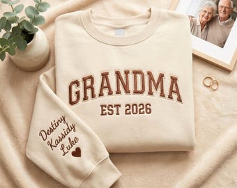 May include: Cream-colored sweatshirt with the word "GRANDMA" embroidered in brown, along with "EST 2026". The left sleeve has embroidered names and a heart. A framed photo and rings are in the background.
