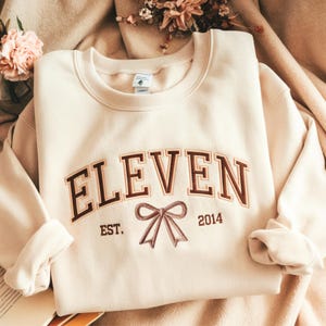 May include: Cream-colored sweatshirt with the word "ELEVEN" in brown, arched lettering. Below the word are the words "EST. 2014" and a bow graphic. The sweatshirt has a crew neck and long sleeves, and is folded on a soft, neutral-toned surface.