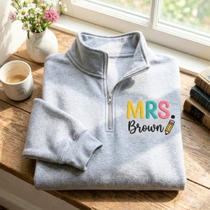 May include: A light gray quarter-zip sweatshirt featuring the embroidered text "MRS. Brown" in colorful letters, with a small yellow pencil graphic next to the name. The garment has a silver-toned metal zipper pull and long sleeves with cuffs.