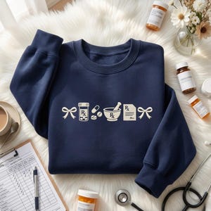 May include: A navy blue crewneck sweatshirt featuring a white graphic print of two bows flanking a prescription bottle, various pills, a mortar and pestle, and a prescription pad with the Rx symbol. The design is centered on the front of the garment.