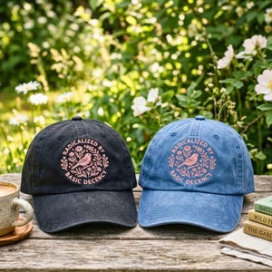 May include: Two baseball caps, one black and one blue, each featuring a circular graphic with a bird, floral elements, and the text "RADICALIZED BY BASIC DECENCY" in pink. The caps have a distressed, washed-out fabric texture and a curved brim.