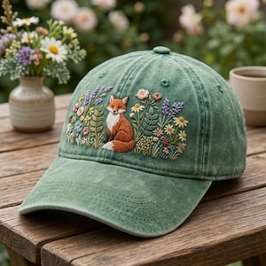 Könnte beinhalten: Eine salbeigrüne Baseballkappe mit einer detaillierten Stickerei eines Rotfuchses, der inmitten bunter Wildblumen wie Gänseblümchen und Lavendel sitzt, zentriert auf der Vorderseite mit einem gebogenen Schirm und gestickten Ösen.