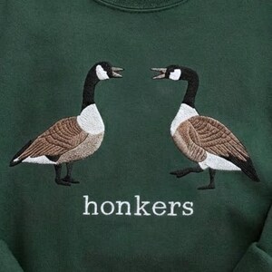 May include: Dark green sweatshirt featuring an embroidered design of two geese facing each other. The geese have brown, white, and black details. The word "honkers" is embroidered in white below the geese.