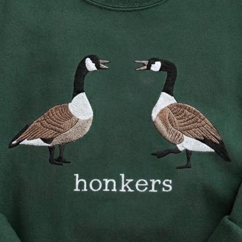 May include: Dark green sweatshirt featuring an embroidered design of two geese facing each other. The geese have brown, white, and black details. The word "honkers" is embroidered in white below the geese.