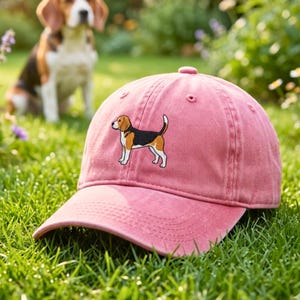 Könnte beinhalten: Staubrosa Baseballkappe mit einem gestickten Beagle-Hund-Design. Die Kappe hat einen gebogenen Schirm und eine Konstruktion aus sechs Paneelen. Der Beagle ist in Schwarz, Braun und Weiß dargestellt. Die Kappe liegt auf grünem Gras.