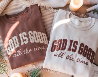 Könnte beinhalten: Zwei Rundhals-Sweatshirts, eines in Braun und eines in Creme, die jeweils den Text "GOD IS GOOD all the time" in einer Schriftart im College-Stil tragen. Das braune Sweatshirt hat cremefarbene Buchstaben, das cremefarbene braune.