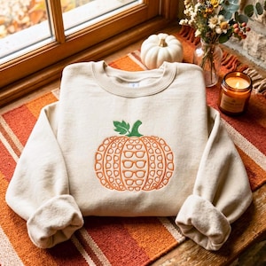 May include: A beige crewneck sweatshirt featuring an embroidered orange pumpkin graphic on the center chest. The pumpkin design is composed of multiple small, stacked eyeglass frames in orange outline with a solid green stem at the top.