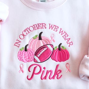 May include: A light pink sweatshirt featuring an embroidered design of three pumpkins, a football, and two breast cancer awareness ribbons. The text reads IN OCTOBER WE WEAR Pink in a mix of block and script lettering with pink thread.