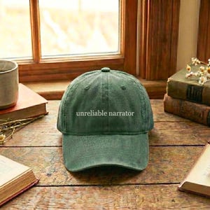 May include: A green baseball cap with the text "unreliable narrator" in white. The cap is sitting on a wooden surface near a window. The cap has a curved brim and a rounded top.