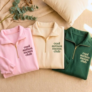 May include: Three quarter-zip sweatshirts in pink, cream, and green. Each sweatshirt has the text "cool autism moms club" embroidered on the chest. The sweatshirts are made of a soft material and have gold-colored zippers.