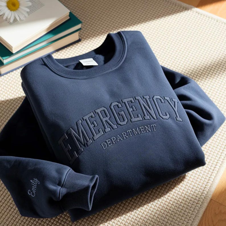 K&ouml;nnte beinhalten: Marineblaues Sweatshirt mit grau gesticktem Schriftzug "EMERGENCY DEPARTMENT" auf der Brust. Am &Auml;rmelbund ist der Name "Emily" in passendem Grauton gestickt. Das Sweatshirt liegt gefaltet auf einem neutralfarbenen Teppich.