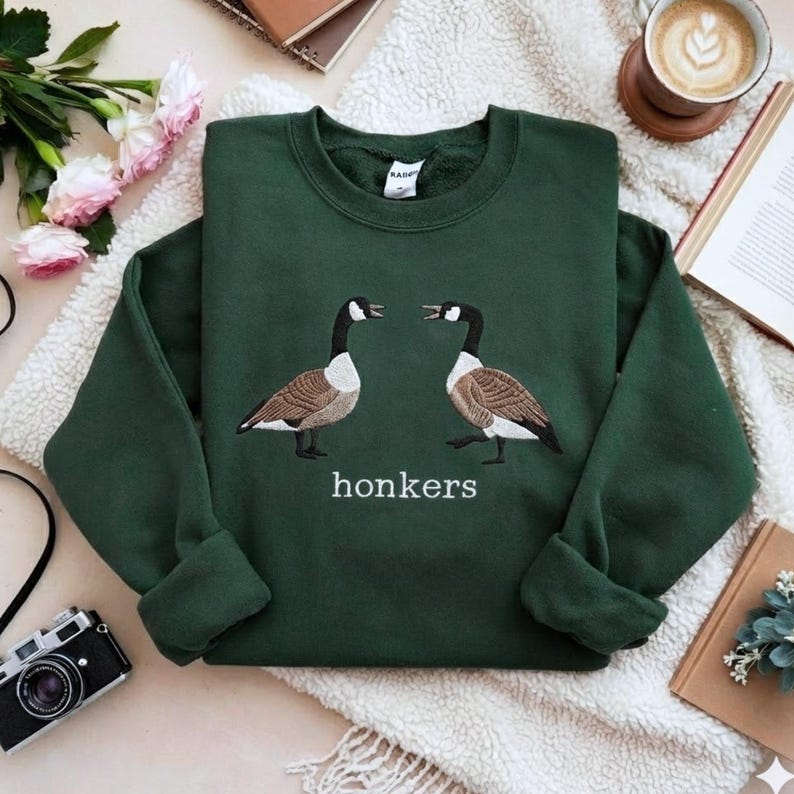 May include: A dark green sweatshirt featuring an embroidered design of two geese facing each other. The word "honkers" is embroidered below the geese in white. The sweatshirt is made of a soft material.