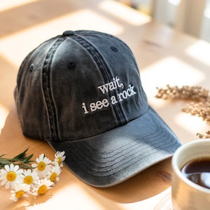 Könnte beinhalten: Dunkelgraue Baseballkappe mit dem gestickten Text "wait, i see a rock" in Weiß. Die Kappe liegt auf einer Holzoberfläche, mit einer Tasse Kaffee und Gänseblümchen im Hintergrund.