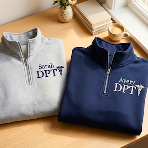 May include: Two quarter-zip pullovers, one light gray and one navy blue, are displayed on a wooden surface. Each features a name, the letters "DPT", and a medical symbol. The gray pullover is labeled "Sarah", and the blue one is labeled "Avery".