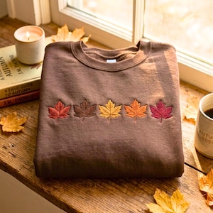 May include: A dark brown crewneck sweatshirt featuring a horizontal row of five embroidered maple leaves across the chest. The leaves are stitched in varying shades of red, brown, yellow, orange, and magenta, creating a colourful seasonal pattern.