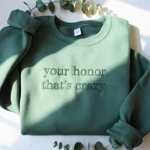 May include: A sage green sweatshirt with the embroidered phrase "your honor, that's crazy." The crew neck sweatshirt has long sleeves with ribbed cuffs. The text is in a matching green thread.