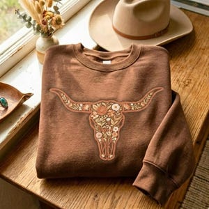 May include: A brown sweatshirt featuring a floral bull skull design. The bull skull is filled with flowers in shades of cream, peach, and green. The sweatshirt is folded on a wooden surface.