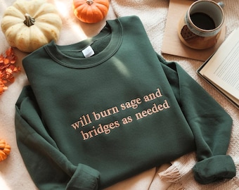 May include: Dark green sweatshirt with the text "will burn sage and bridges as needed" in a light orange color. The image includes small pumpkins, a mug, and a book, suggesting a fall theme.