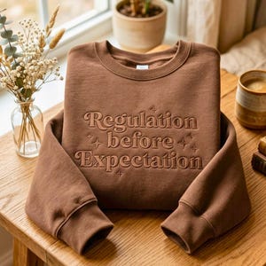 Puede incluir: Una sudadera de cuello redondo en color marrón con el texto bordado "Regulation before Expectation" en hilo marrón a juego. El diseño incluye pequeños detalles de estrellas bordadas que rodean las letras en la parte frontal.