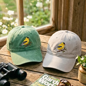 Puede incluir: Dos gorras de béisbol, una verde y otra gris, sobre una superficie de madera. Cada gorra presenta un bordado de un pájaro amarillo posado en una rama. También se ven unos prismáticos y una revista de jardinería.