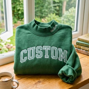 May include: A forest green sweatshirt with a mock neck collar featuring the word custom embroidered in white script. The front displays the word CUSTOM in large, white-bordered, green and white gingham checkered block letters.