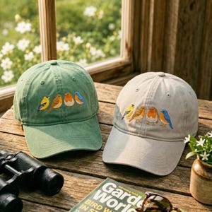 Puede incluir: Dos gorras de béisbol, una en verde salvia y otra en gris claro, cada una con un diseño bordado de cuatro pájaros pequeños en colores amarillo, marrón, naranja y azul. Las gorras tienen una visera curva y una corona estructurada.