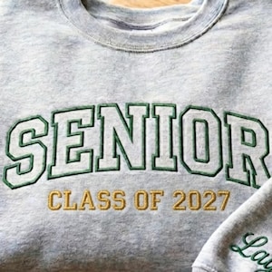 May include: A heather gray sweatshirt with the word "SENIOR" embroidered in green block letters with a green outline. Below the word "SENIOR" is the text "CLASS OF 2027" in gold thread. The sweatshirt is a crew neck style.