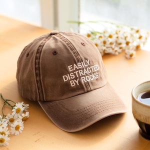 May include: Brown baseball cap with the text "EASILY DISTRACTED BY ROCKS" embroidered in white. The cap is made of a soft, worn material and has a curved brim. The cap is sitting on a wooden surface.