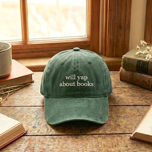 May include: A green baseball cap with the text "will yap about books" in white. The hat is sitting on a wooden surface with books and a window in the background.