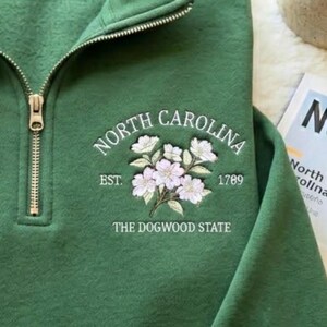 Może przedstawiać: Zielona bluza z dekoltem zapinanym na zamek i haftowanym kwiatowym wzorem. Projekt zawiera napisy "North Carolina", "Est. 1789" i "The Dogwood State". Bluza ma złoty zamek.