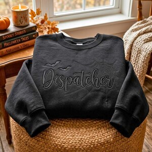 Puede incluir: Una sudadera negra de manga larga con el texto bordado Dispatcher en letra cursiva. El diseño incluye una telaraña, una pequeña araña y dos murciélagos volando, cosidos con hilo negro a tono en la parte frontal del pecho.