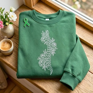 Könnte beinhalten: Ein waldgrüner Rundhals-Pullover mit einer weißen Strichzeichnung einer menschlichen Wirbelsäule, die mit blühenden Rosen verflochten ist. Eine kleine grüne Anstecknadel in Form einer Schleife ist im oberen linken Brustbereich befestigt.