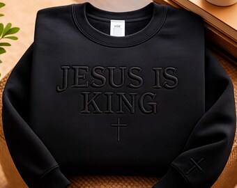 May include: Black crewneck sweatshirt with the embossed text "JESUS IS KING" and a cross. The sweatshirt has a small cross detail on the left sleeve. The VGE brand label is visible on the neckline.