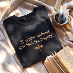 May include: Black sweatshirt with gold text reading "A reader without their book is a tragedy" and a dragon graphic. A cup of coffee and an open book are also in the image.