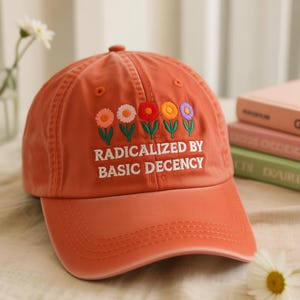 May include: A coral-colored baseball cap with embroidered flowers and the text "RADICALIZED BY BASIC DECENCY." The cap features a curved brim and a classic six-panel design. The flowers are pink, white, red, yellow, and purple.