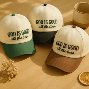 May include: Three baseball caps with the text "GOD IS GOOD all the time". The caps have a cream-coloured crown and a coloured brim in green, dark green, and brown. The caps are on a light brown surface.