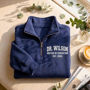 May include: Navy blue pullover sweatshirt with a quarter-zip closure and a white printed design. The text reads "DR. WILSON DOCTOR OF EDUCATION EST. 2026". The sweatshirt is folded and placed on a light-colored surface.