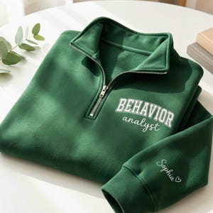 May include: A forest green quarter-zip sweatshirt with the words "BEHAVIOR analyst" in white block letters. The sleeve cuff features the name "Sophia" in cursive with a small heart. The sweatshirt is folded neatly on a white surface.